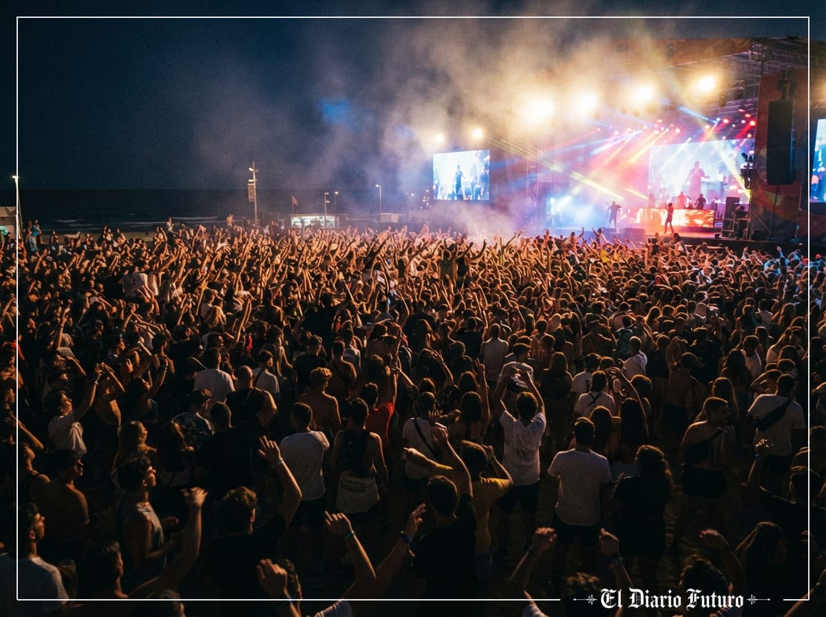 Multitud en un concierto nocturno al aire libre con luces de escenario y los brazos en alto.